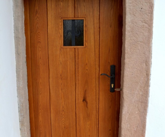 Dveře vstupní fošnové s okénkem a kovanou mříží, jednokřídlé, rámová zárubni v kamenném ostění, dub, drásané, nátěr lazurou.