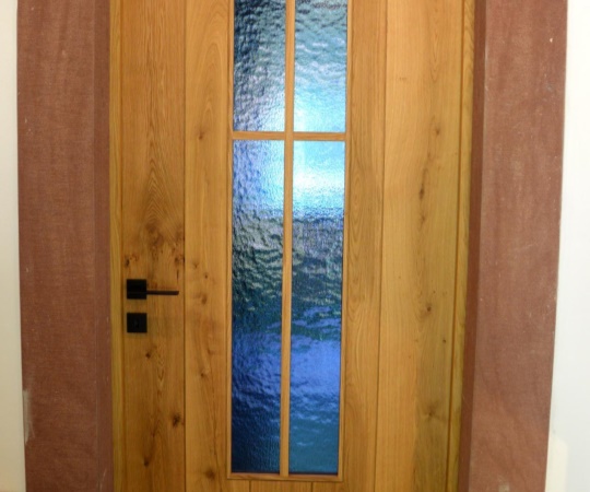 Dveře jednokřídlé fošnové rámová zárubeň v kamenném ostění. Masiv dub, drásané, olejované. Prosklené okénko s křížem.